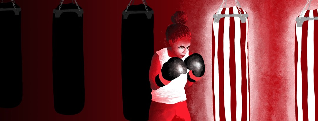 A boxer stands in front of several punching bags that are dark and receding into the background, and behind a punching bag that is lit up and in full color, ready for her to take on this next obstacle.