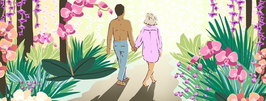A shirtless man holds hands with a woman wearing only a men's dress shirt walk down a brightly lit path lined with orchids and other florals and greenery.