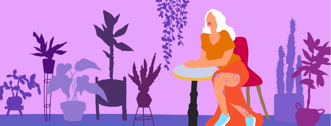 A woman seated at a table surrounded by house plants is wearing a long dress that partially reveals her legs which are covered in purple veins.