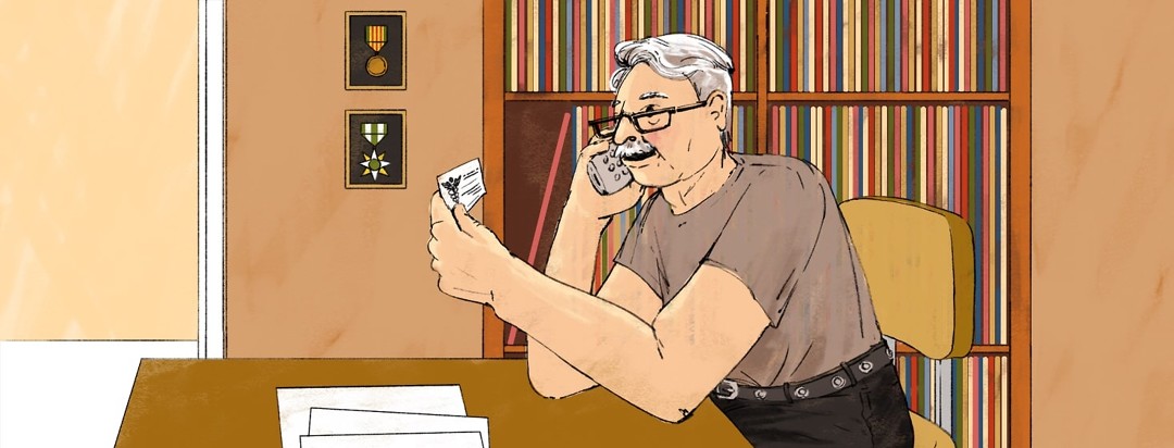 An older man talks on a telephone while looking at a doctor's business card. Behind him on the wall are stacks of records on shelves next to two war medals hanging on the wall.